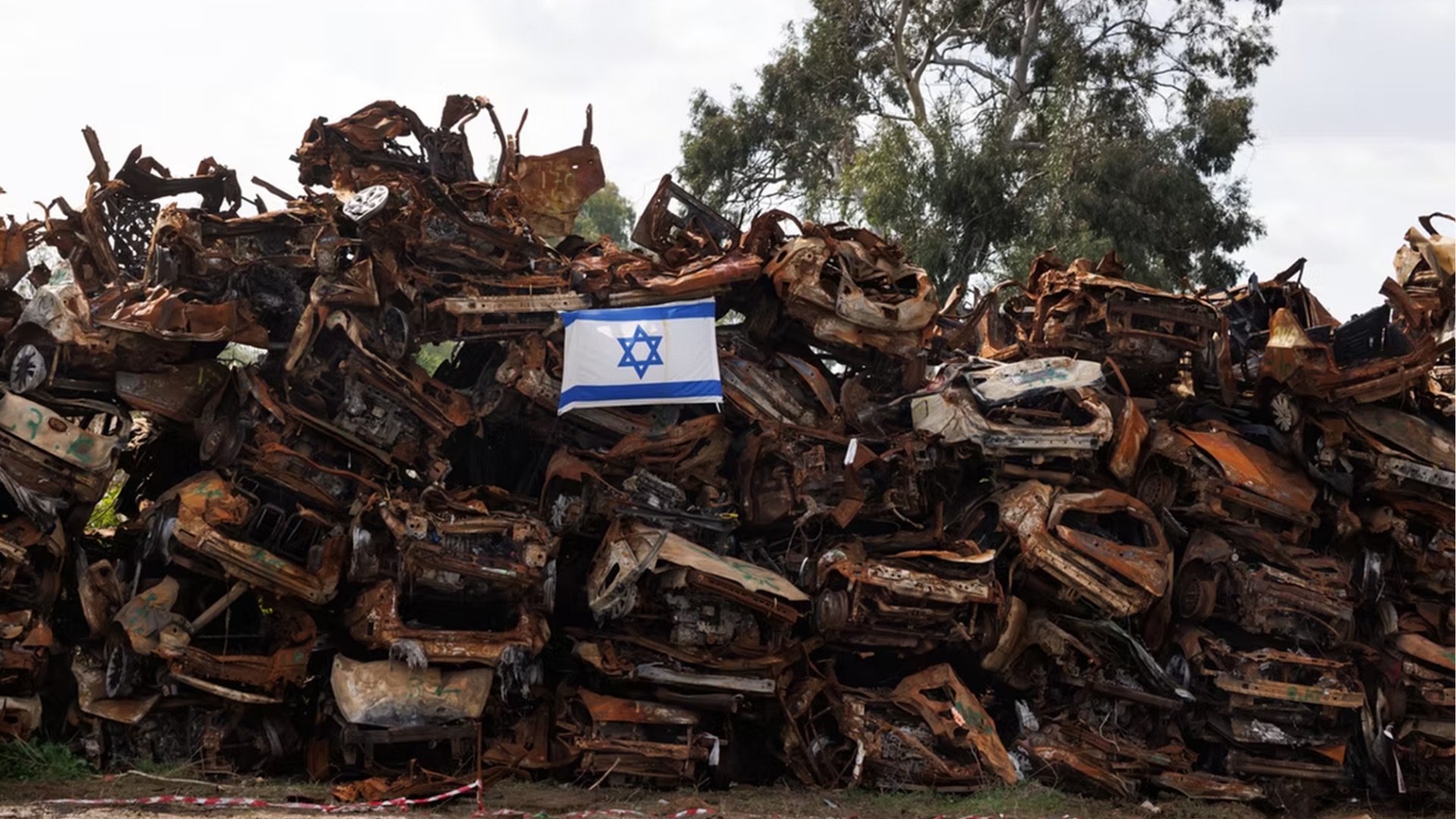 Cientos de autos quemados y perforados, apilados en un terreno. Encima, una bandera de Israel.